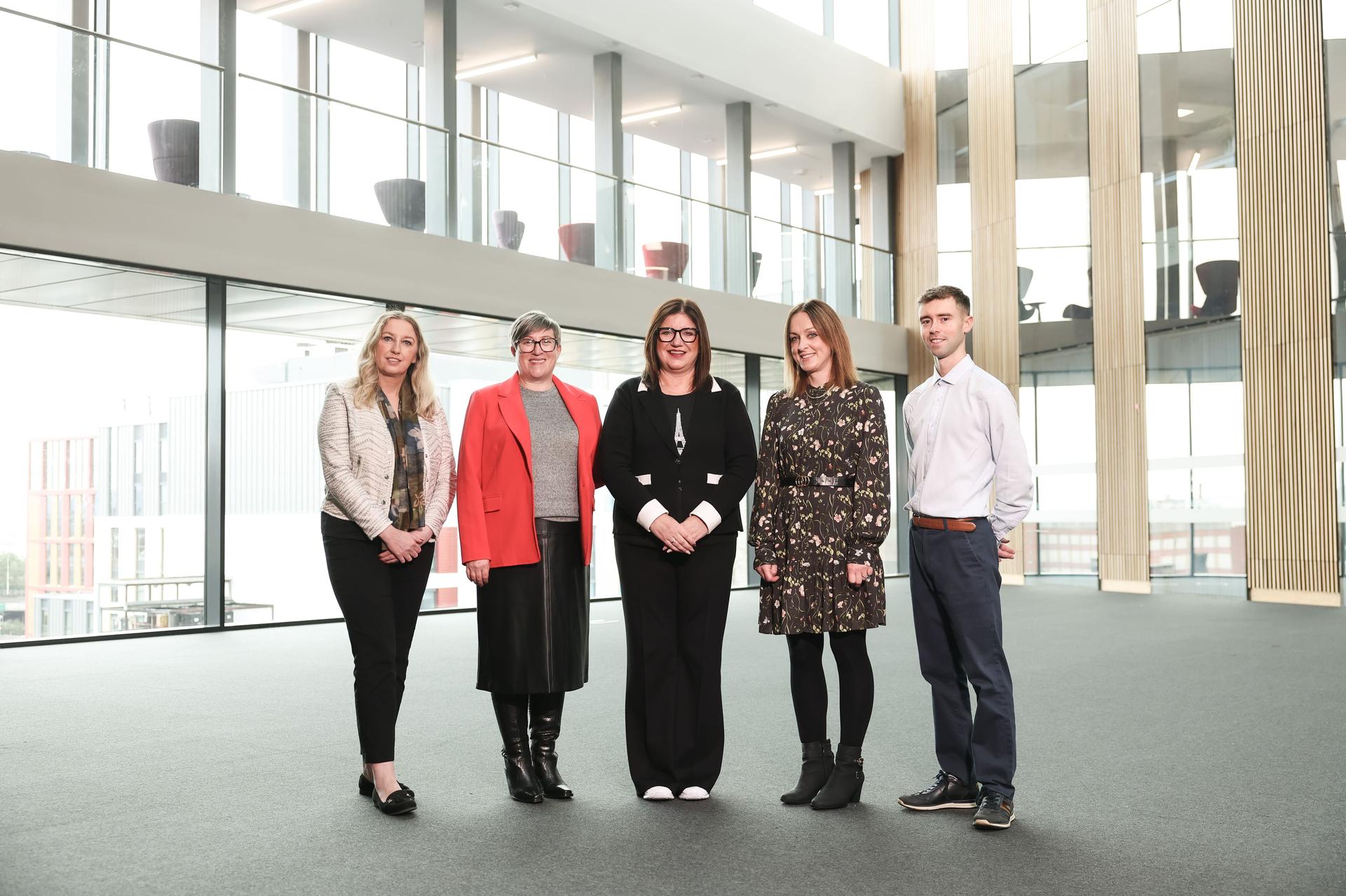 Five people standing in a modern, spacious atrium with large windows and high ceilings. They are dressed in professional attire, conveying a formal tone.